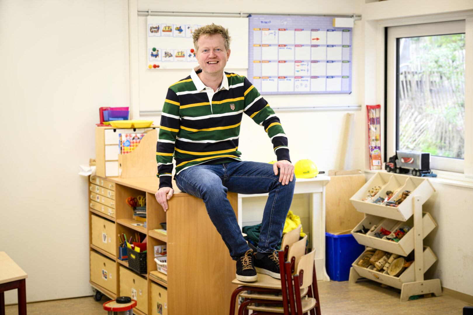 Meestr Mark zittend op een kastje met voeten op een stapel kinderstoeltjes in een klaslokaal,