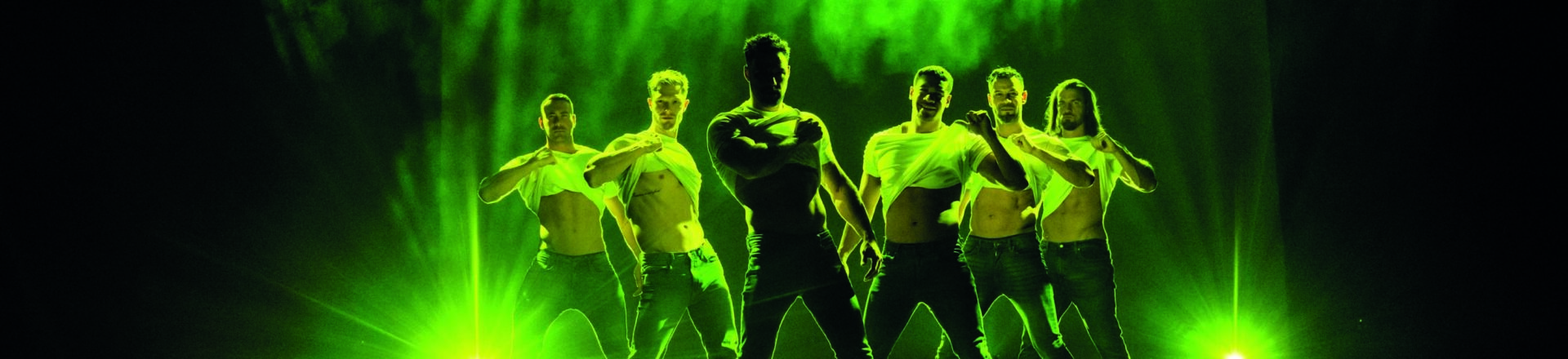 Op het podium staan in groen licht 5 mannelijke dansers met hun T-shirt omhoog.